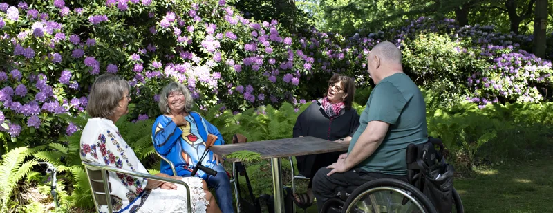 Fire personer sidder ved et bord og taler ude i naturen.
